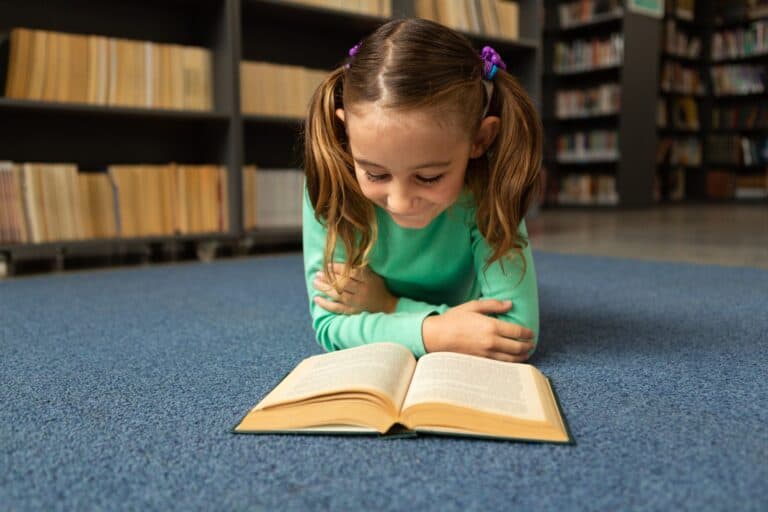 Schoolgirl lying on floor and reading book in library at elementary school